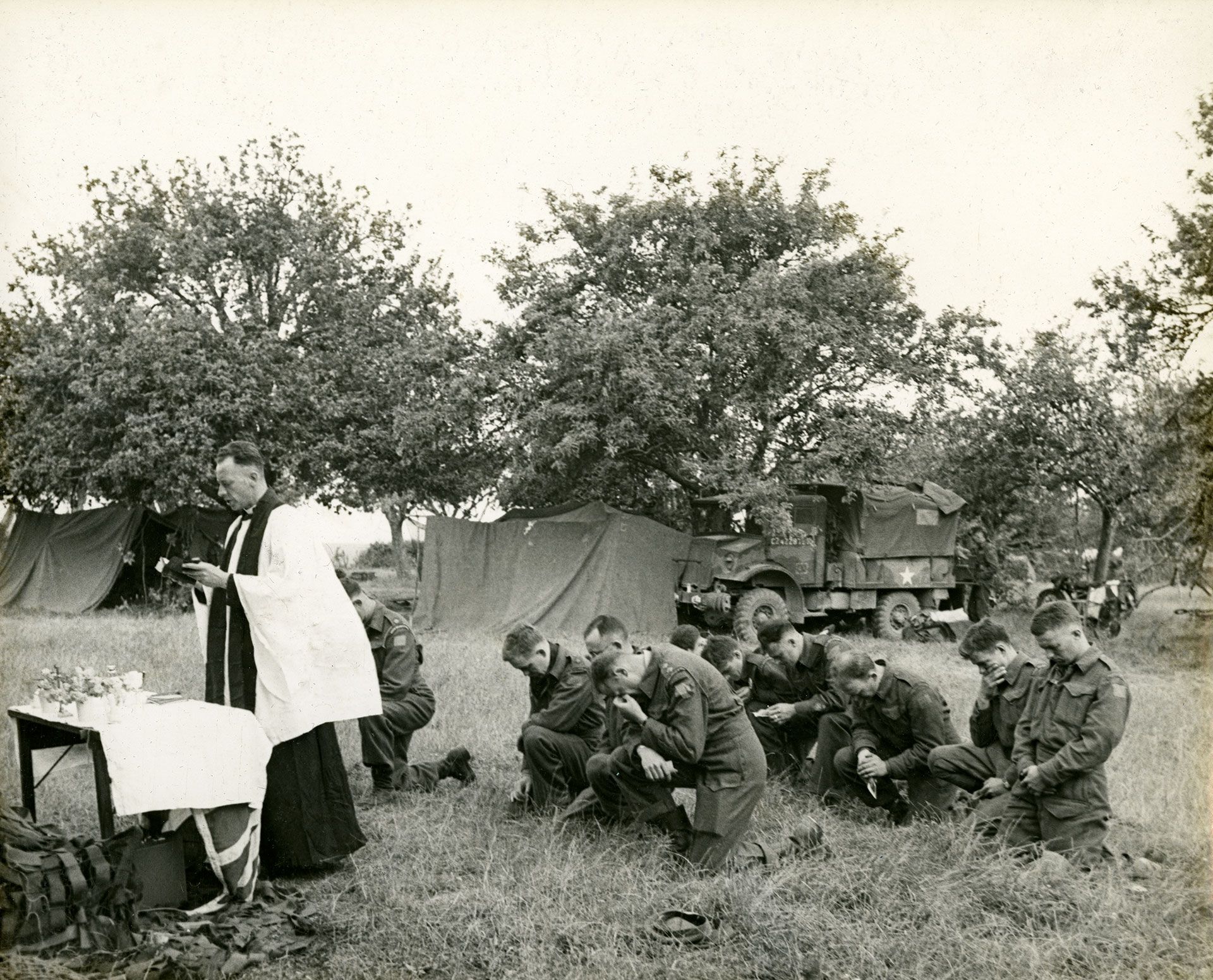 Photo en noir et blanc d’une messe en plein air. Un aumônier, à gauche, tient une bible au-dessus d’une petite table couverte d’un tissu blanc. Deux rangées de soldats sont à genoux derrière lui, dans l’herbe haute, et à l’arrière-plan, il y a des arbres, des tentes et des véhicules militaires. 