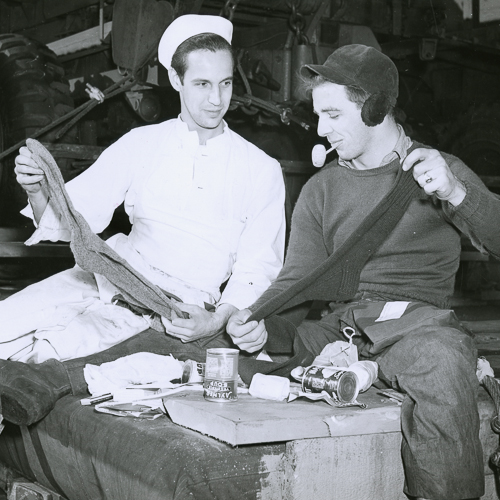 Deux marins assis ensemble tiennent en place deux chaussettes tricotées à la main.