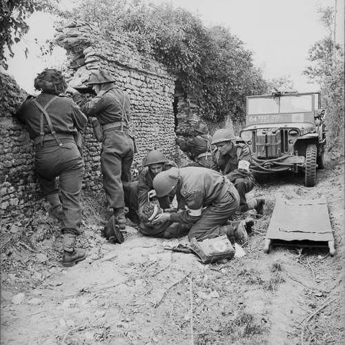 Trois soldats soignent un soldat blessé. Trois autres soldats défendent la position, se tenant derrière un mur de pierre.