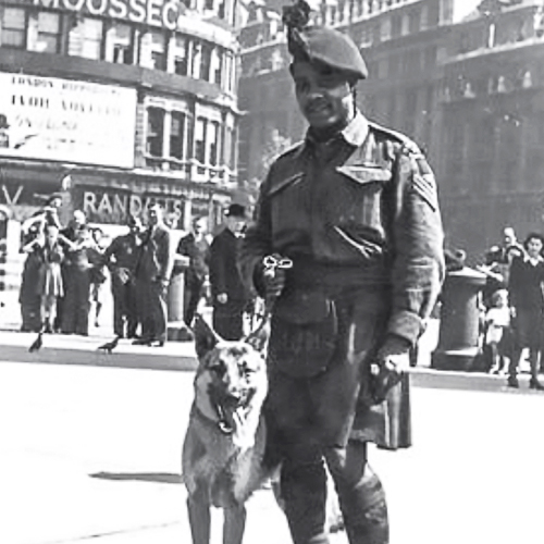 Un soldat afro-canadien portant sa veste de tenue de combat et un kilt pose avec son chien sur une rue à Londres.