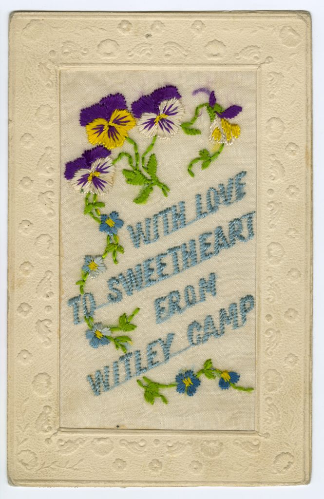 Une carte de couleur crème avec une bordure en relief. Les mots « With love to sweetheart from Witley Camp » (À la bien-aimée avec amour, du camp Witley) sont brodés en fil bleu clair et sont accompagnés de fleurs bleues, violettes et jaunes avec des tiges et des feuilles vertes.