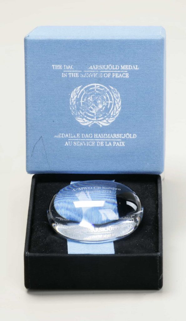 Objet en cristal clair en forme de sphère légèrement aplatie, orné d’une gravure, dans un écrin portant la mention « The Dag Hammarskjöld Medal/In the Service of Peace/Médaille Dag Hammarskjöld/Au service de la paix » sur le couvercle.