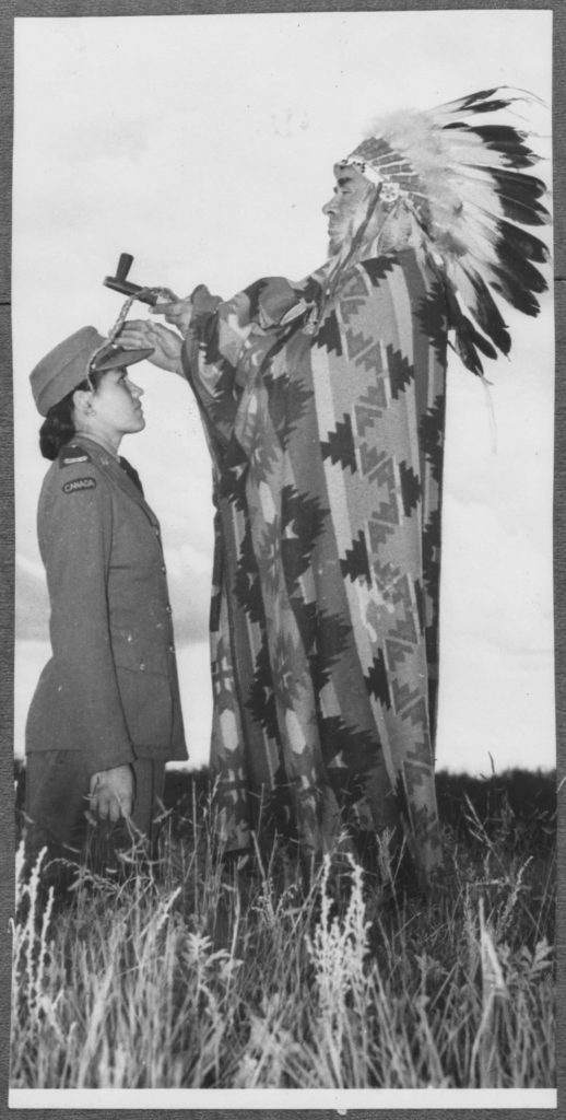 Une femme autochtone en uniforme est agenouillée dans un champ, devant un Autochtone dont les vêtements et les objets de cérémonie sont mal assortis.