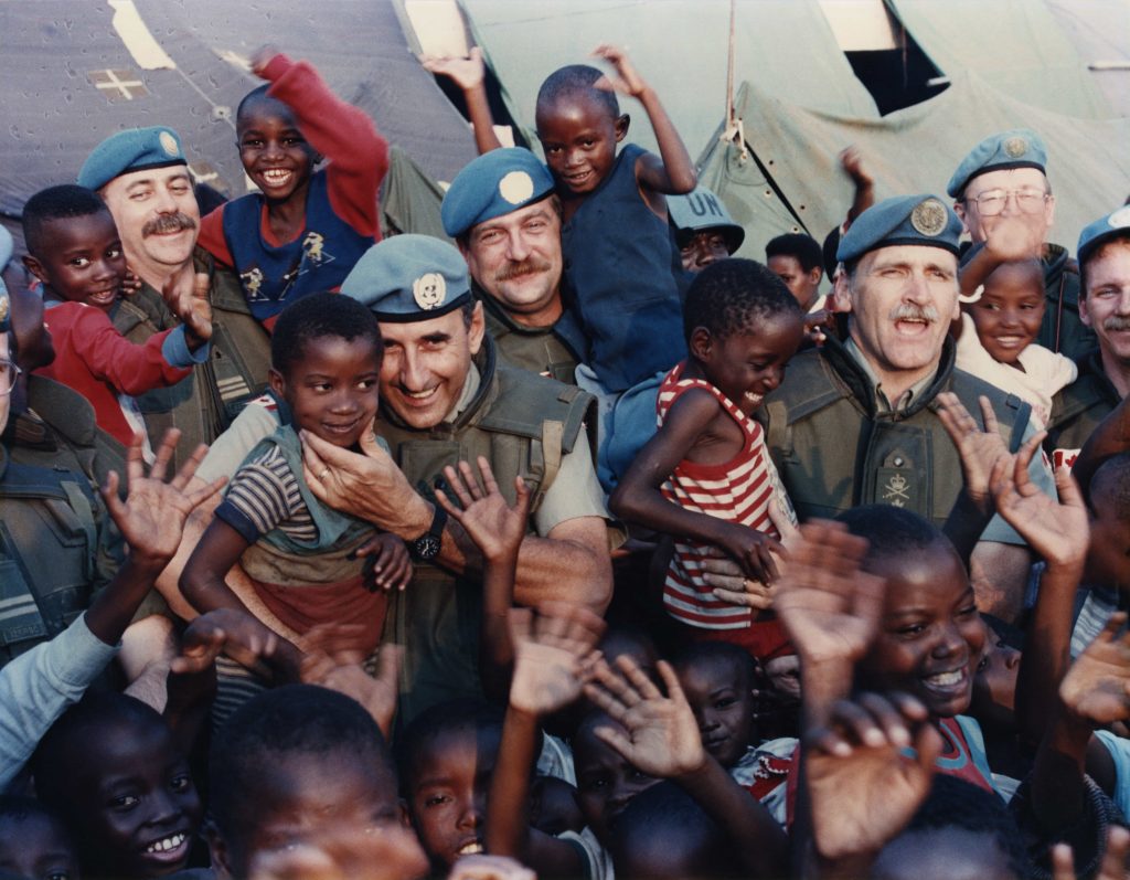 Une foule composée d’enfants noirs et d’hommes blancs portant l’uniforme des Casques bleus sourient et saluent pour la photo.