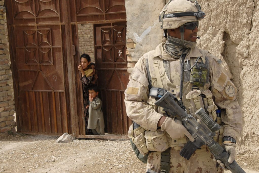 Un homme en uniforme tenant un fusil surveille les environs de l’endroit où il est posté; des enfants derrière lui jettent un coup d’œil par une grande porte en bois.