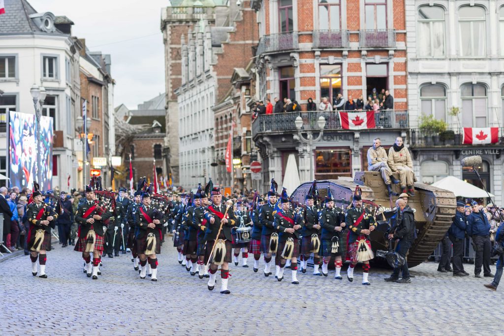 Des hommes portant des uniformes de cérémonie défilent dans une rue de Mons, le long de bâtiments décorés de drapeaux canadiens. Deux hommes sont assis sur un char d’assaut de l’époque de la Première Guerre mondiale.