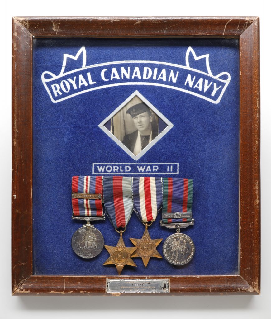 Une photographie en noir et blanc de John Laing en uniforme et ses médailles disposées en dessous. Les objets sont présentés dans un cadre en bois sombre et mis en valeur sur un fond bleu. Les mots « Royal Canadian Navy » (Marine royale du Canada) et « World War II » (Seconde Guerre mondiale) figurent en haut et au centre respectivement, dans le cadre.