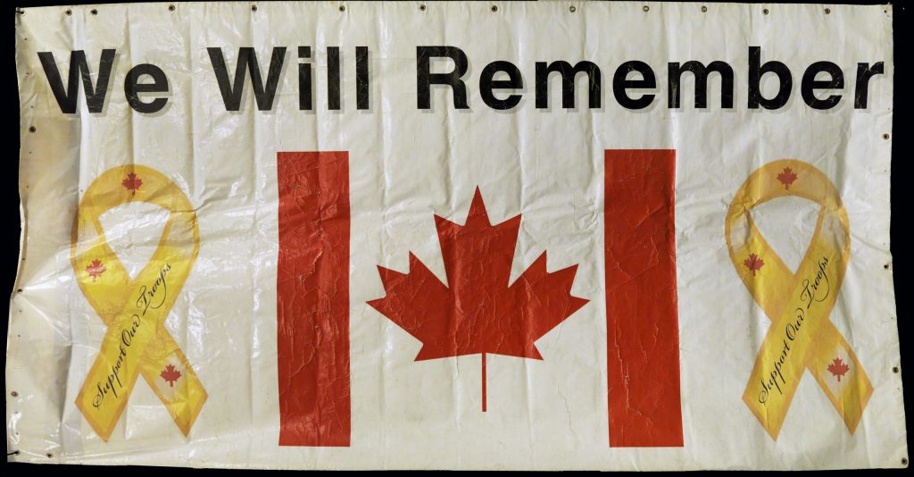 Une bannière blanche usée avec des œillets métalliques sur trois côtés. Le drapeau canadien au milieu est encadré de rubans jaunes portant les mots « Support Our Troops » (Appuyons nos troupes). Les mots « We Will Remember » (Nous nous souviendrons) sont imprimés en gros caractères en haut de la bannière.