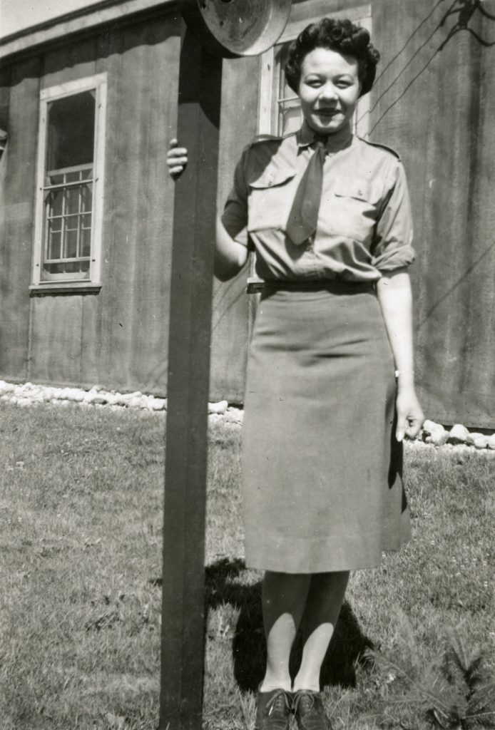 Minnie Gray en uniforme, debout à côté d’un poteau en bois, avec une cabane en bois et une pelouse à l’arrière-plan.