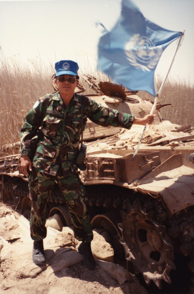 Ted Itani hisse un drapeau bleu et blanc des Nations Unies. Il porte un uniforme camouflé, une casquette bleue et des lunettes de soleil. À l’arrière-plan, il y a un char détruit.