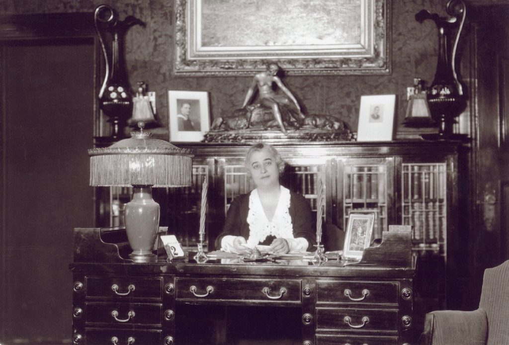 Lillian Bilsky Freiman assise à son bureau, entourée d’une grande lampe, de plusieurs cadres, d’une statue d’une figure allongée et de deux grands vases.