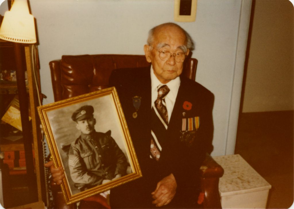 Masumi Mitsui à un âge avancé, portant l’uniforme de la Légion royale canadienne. Il tient une photo encadrée de lui-même, en noir et blanc, prise quand il était un jeune soldat.