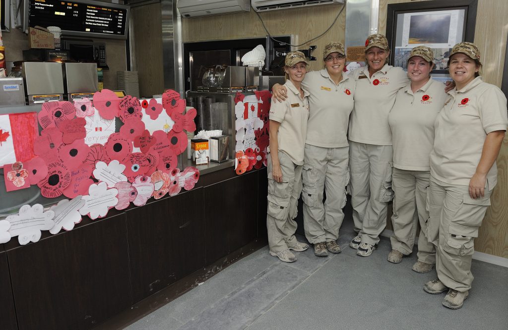 Dans une pièce décorée de coquelicots en papier rouge et de lettres, cinq travailleurs portant l’uniforme de Tim Hortons et un coquelicot sourient et se tiennent les bras autour les uns des autres.