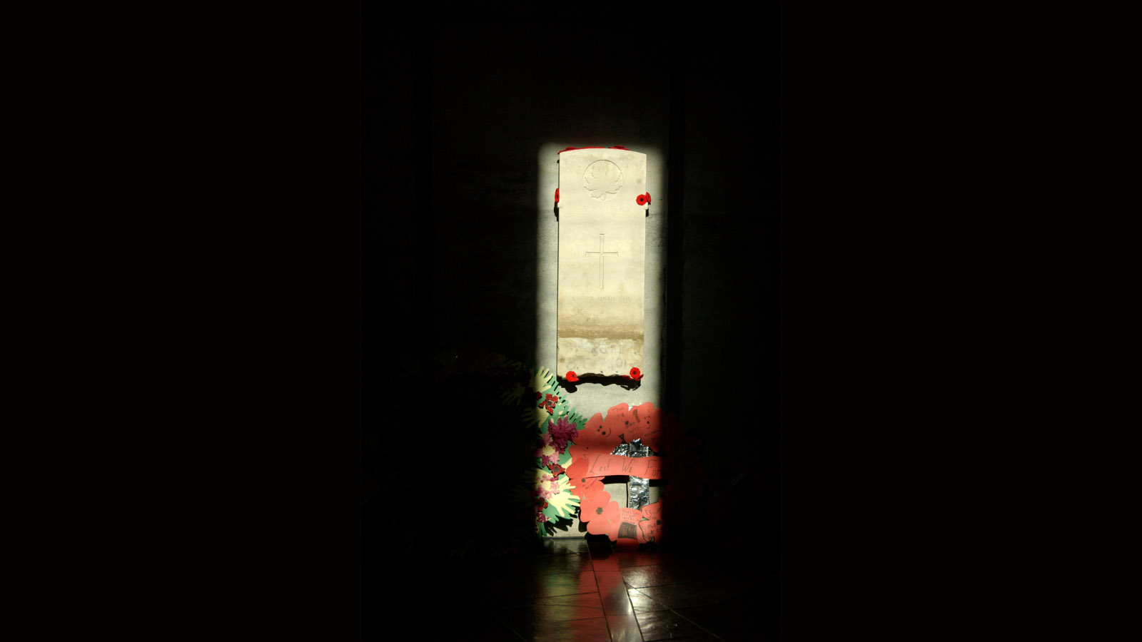 Headstone in pitch black, illuminated by the sun . Paper poppy wreath below.