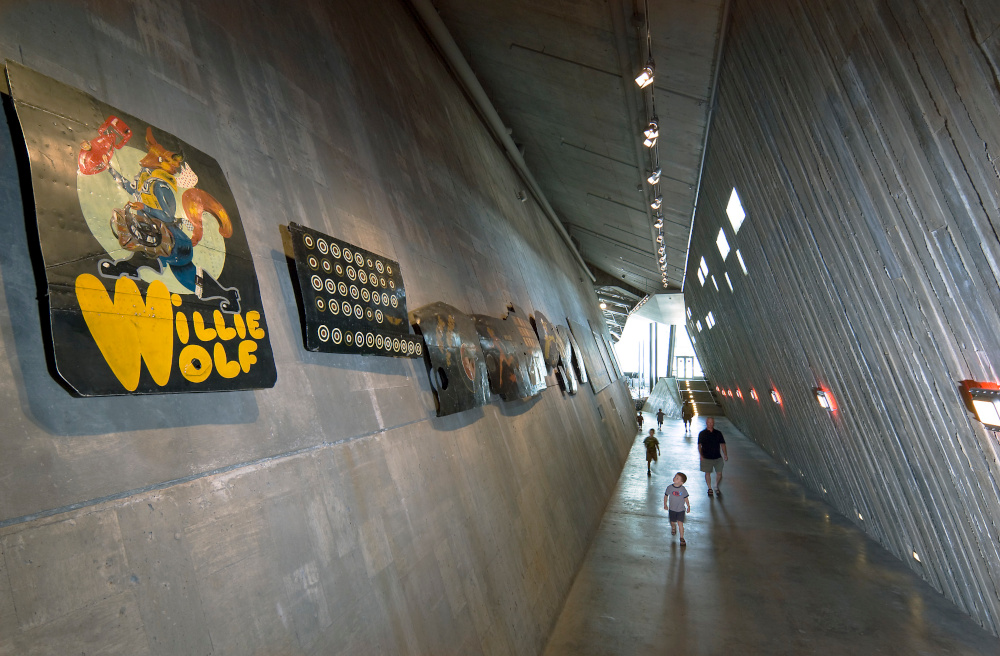 Un long et haut corridor en béton, aux murs fortement inclinés et au toit en angle, mène à une grande pièce ensoleillée au loin. Des tableaux sont accrochés en hauteur au mur de gauche.