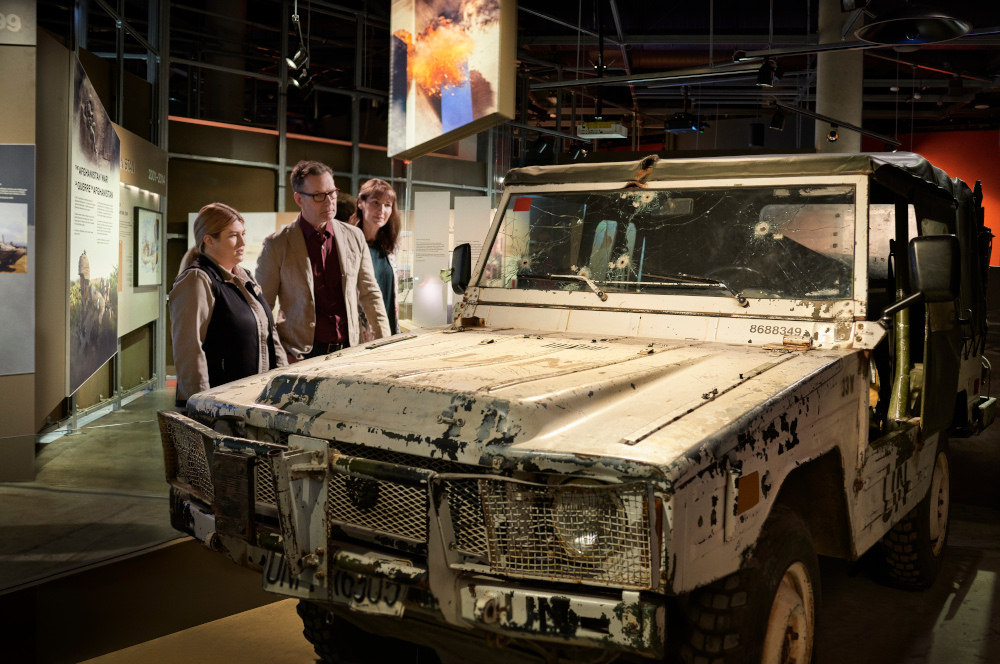 Trois personnes se trouvent dans une galerie du Musée et regardent une Jeep blanche abimée dont le pare-brise est criblé de trous de balle.