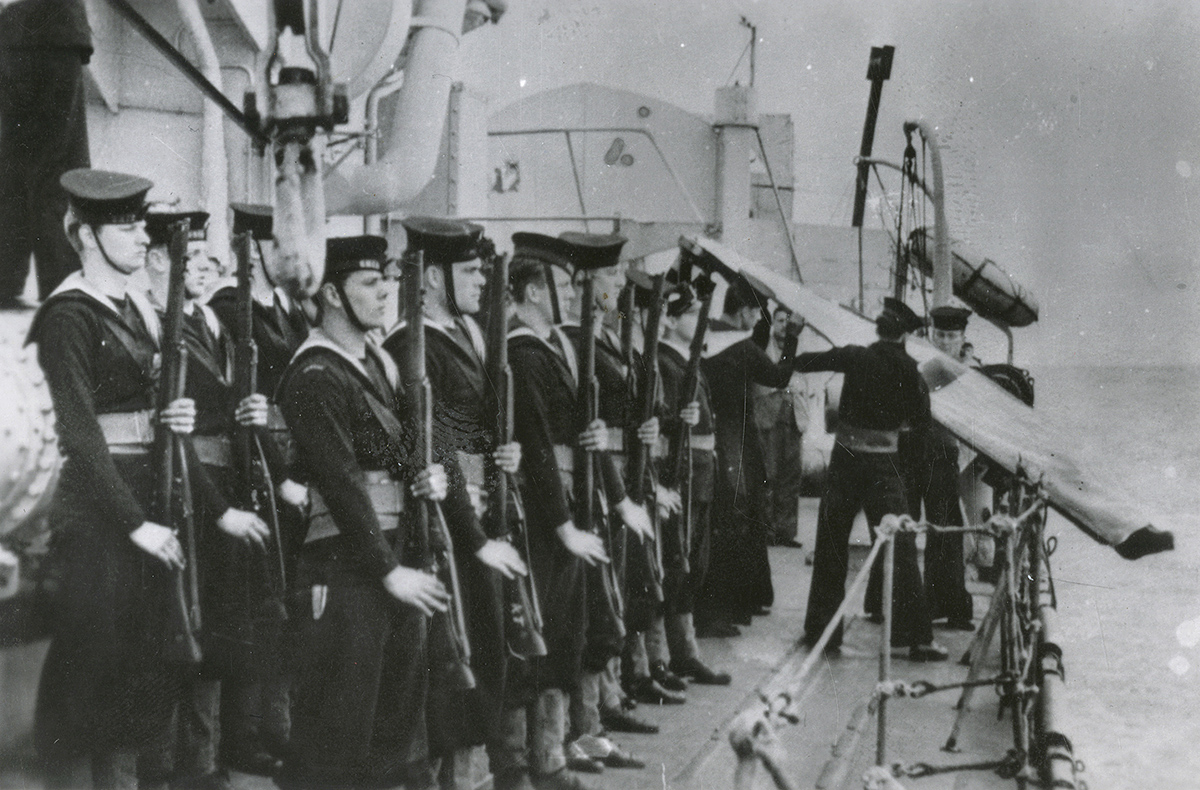Photographie de marins tenant des fusils, en formation sur le pont d’un navire. Deux marins basculent un corps dans un linceul par-dessus le flanc du navire.