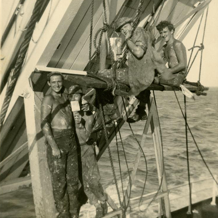 Photographie de quatre marins souriants, couverts d’éclaboussures de peinture alors qu’ils peignent un radeau de sauvetage.