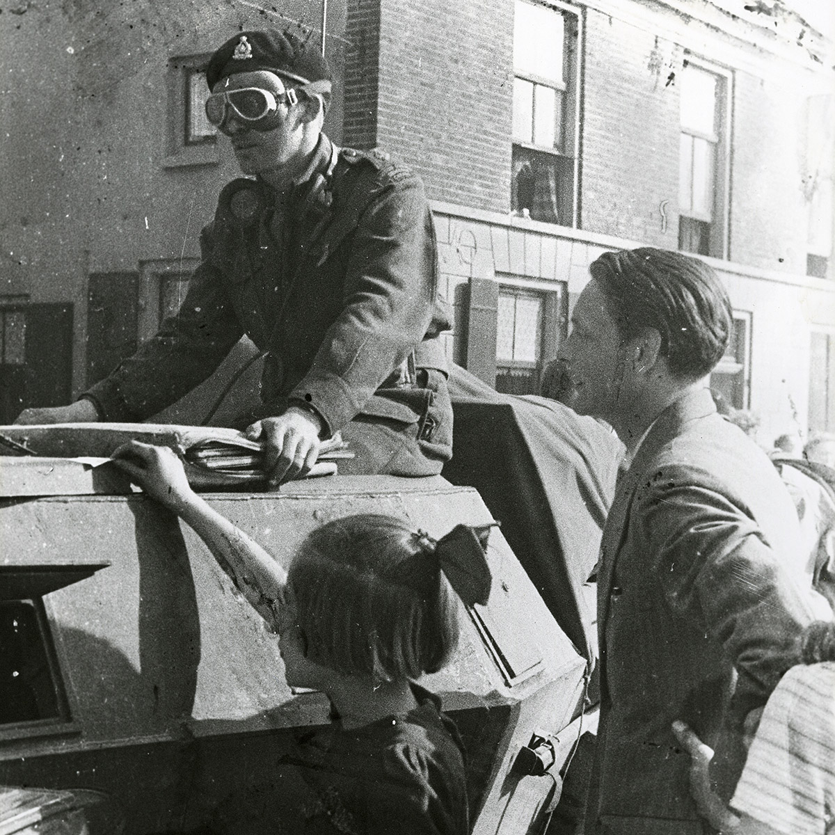 Photographie d’un officier portant des lunettes et un béret, assis sur un véhicule blindé. Un adulte et une enfant se tiennent sur le côté du véhicule.