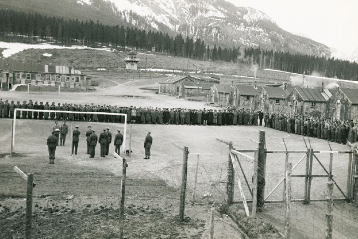Photo d’un camp d’internement avec des personnes détenues se tenant debout dans un champ. Des cabanes sont au second plan et des montagnes sont en arrière-plan.