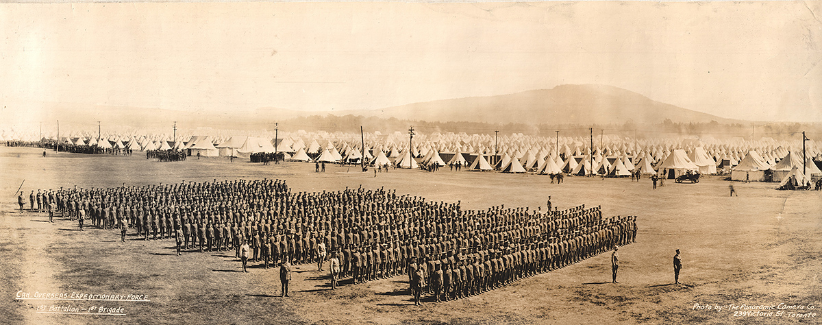Photographie d’un bataillon en formation de parade au premier plan et d’un camp avec de nombreuses tentes à l’arrière-plan.