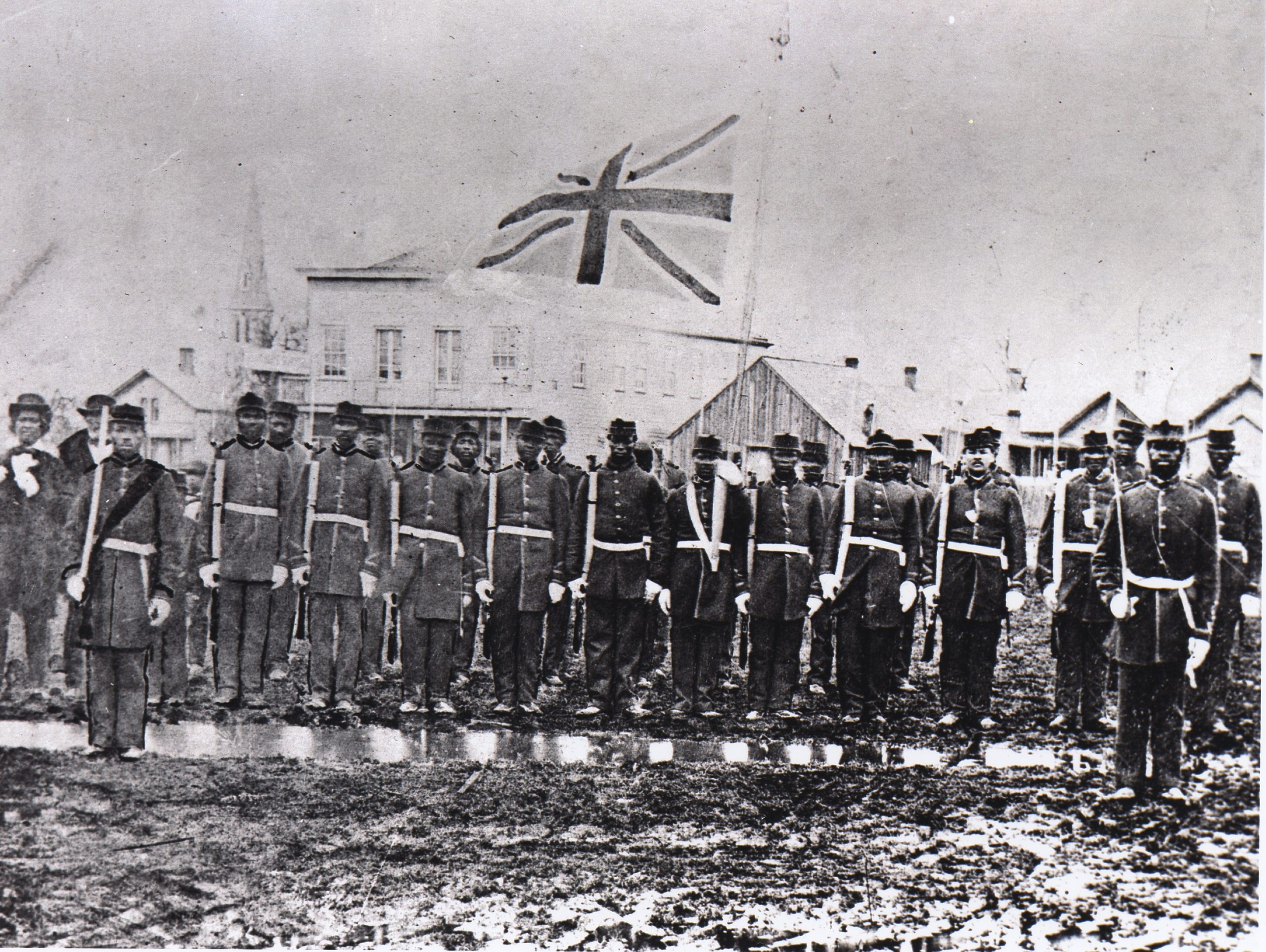 Black and white photo of black soldiers in a line. The Union Jack  flag fliest above them.