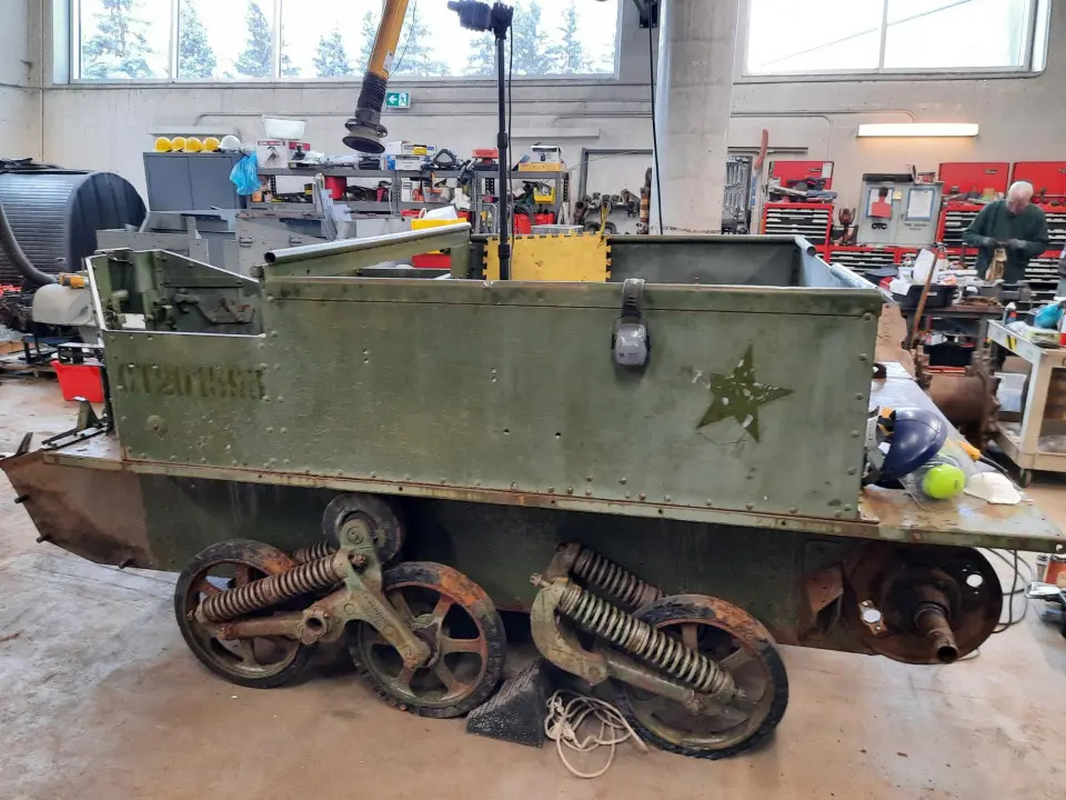 Le même véhicule blindé dans un grand garage rempli d’outils. Ses chenilles ont été retirées, de la peinture plus foncée laisse apparaître les endroits où ses marquages ont été effacés, et ses roues sont visiblement rouillées.