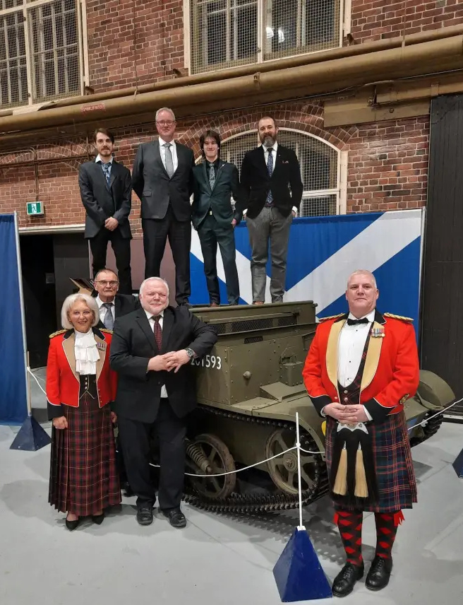 Plusieurs personnes en tenue de cérémonie, dont deux portant des vestes rouge vif et des kilts en tartan, posent debout autour et sur le toit d’un véhicule blindé bas et carré.