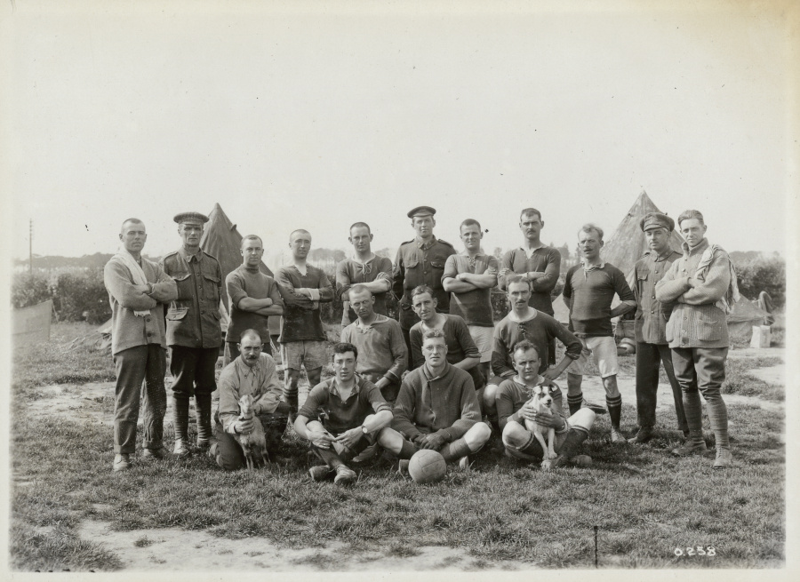 Un groupe de 18 personnes posent en rangées derrière un ballon de soccer. Certaines portent un short et un tee-shirt à manches courtes, d’autres un uniforme militaire. À l’extrême gauche du premier rang, un homme souriant tient une petite chèvre; à l’extrême droite, un homme tient un petit chien sur ses genoux.