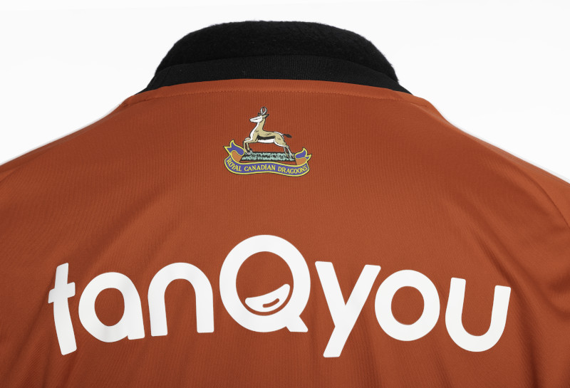 Gros plan sur le milieu du dos d’un tee-shirt de sport à manches courtes, de couleur orange-marron, orné du logo d’une antilope au-dessus d’une banderole portant l’inscription « Royal Canadian Dragoons » et d’un grand texte blanc indiquant « tanQyou ».