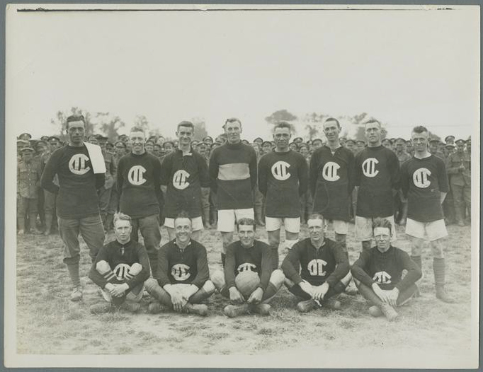 Photo d’équipe en noir et blanc montrant huit personnes à la peau pâle, aux cheveux très courts, debout derrière cinq autres assis par terre. La personne assise au milieu tient un ballon de soccer. La plupart portent un short clair et un tee-shirt foncé arborant un logo bien visible. Une foule importante de personnes en uniforme militaire remplit l’arrière-plan.
