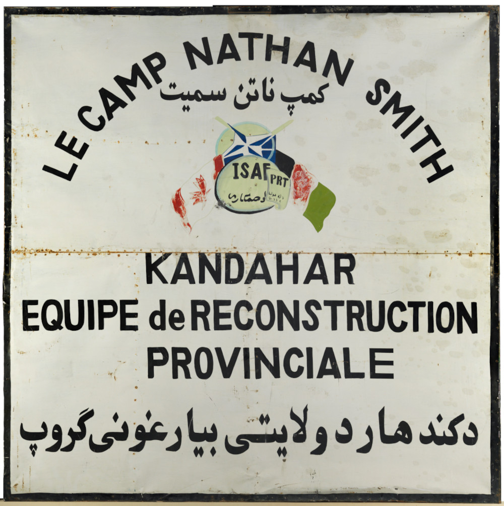 Un panneau carré blanc sur lequel figure un dessin des drapeaux du Canada et de l’Afghanistan, très usé, ainsi qu’une inscription en grand en français et en arabe : « LE CAMP NATHAN SMITH KANDAHAR ÉQUIPE DE RECONSTRUCTION PROVINCIALE. »]