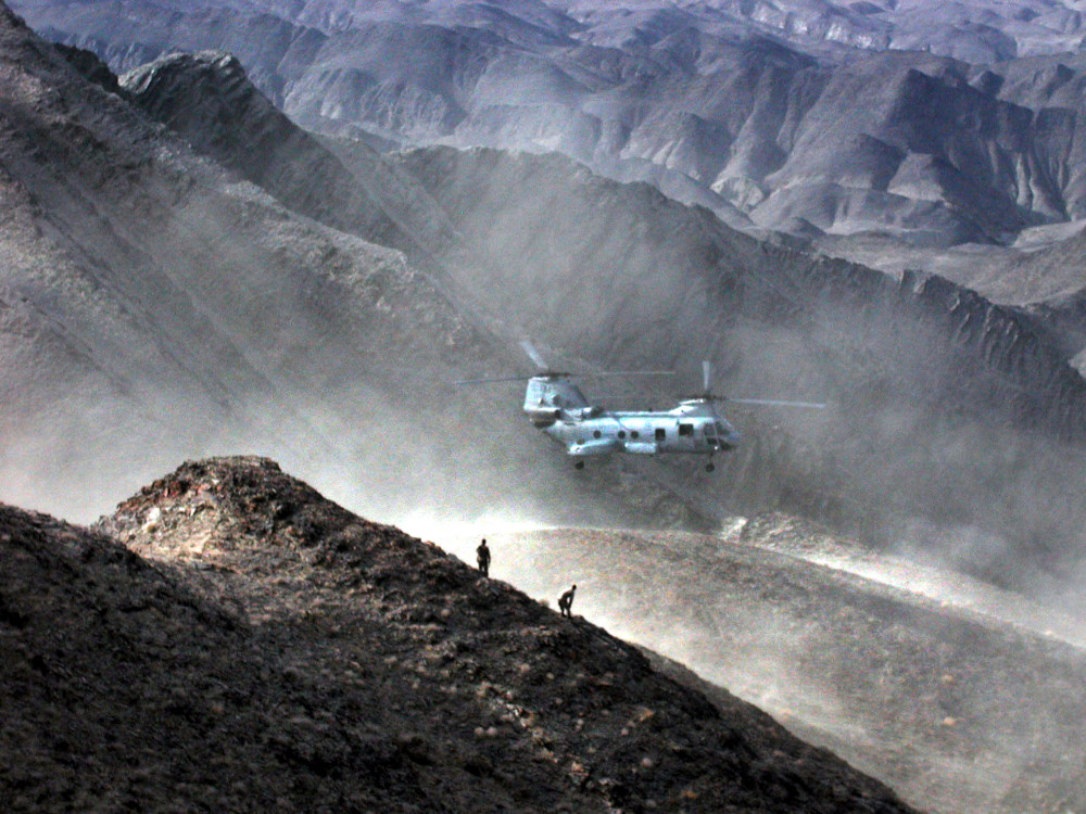 Un gros hélicoptère à deux rotors en vol stationnaire au milieu d’un paysage montagneux, où l’on distingue deux minuscules silhouettes humaines sur la crête d’une montagne.