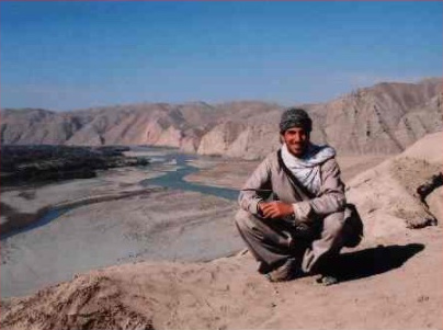 Un homme souriant, à la peau de teint moyen, coiffé d’un chapeau de laine et vêtu d’habits amples de couleur beige, est accroupi sur une crête de montagne peu élevée, face à la caméra, avec une vallée fluviale en arrière-plan.