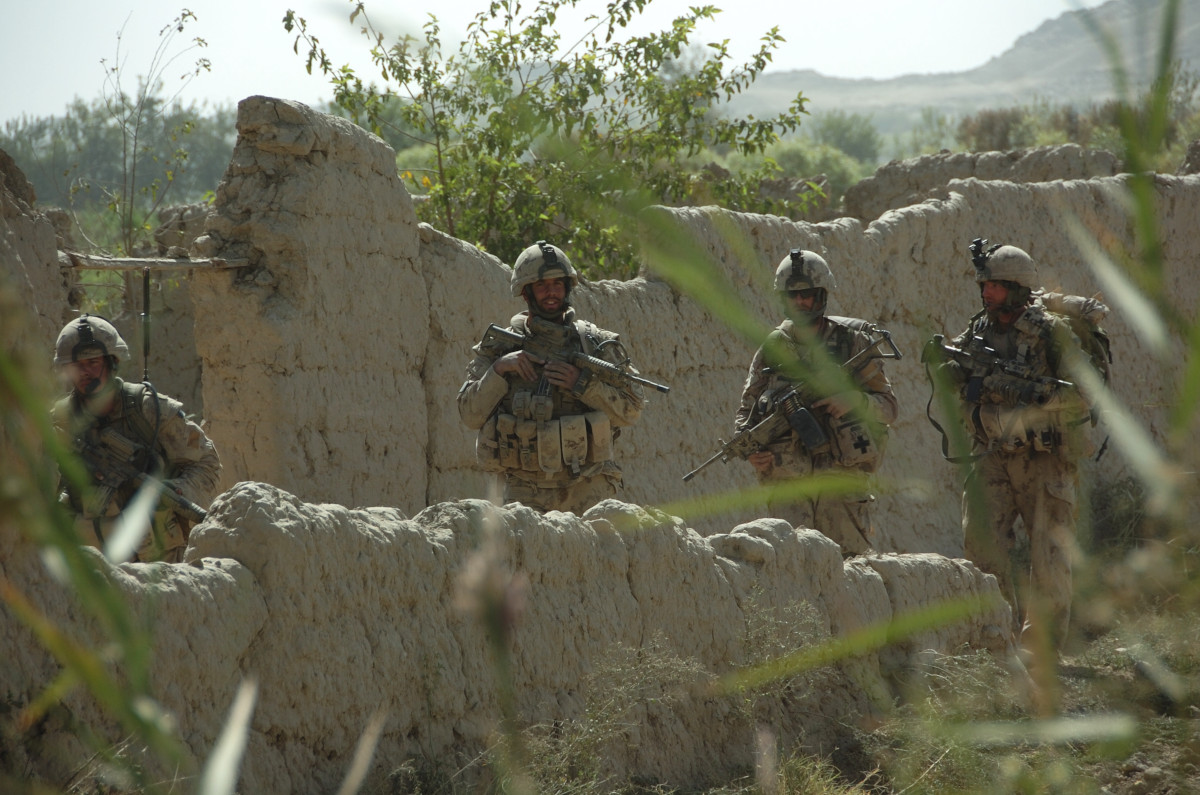 Quatre militaires portant une tenue de camouflage beige, avec leurs armes, et marchant entre des murs de boue, au milieu d’une végétation luxuriante.