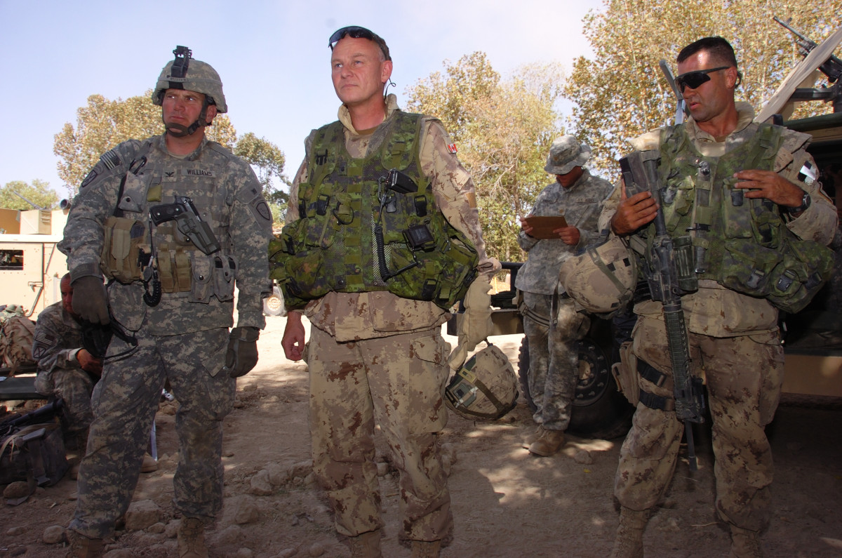 [Un soldat aux cheveux gris courts, vêtu d’une tenue de camouflage beige et d’un gilet vert, entre un soldat en tenue de camouflage gris portant un casque (à gauche) et un soldat aux cheveux foncés très courts, portant des lunettes de soleil, une tenue de camouflage beige, un gilet vert et une arme à feu (à droite).