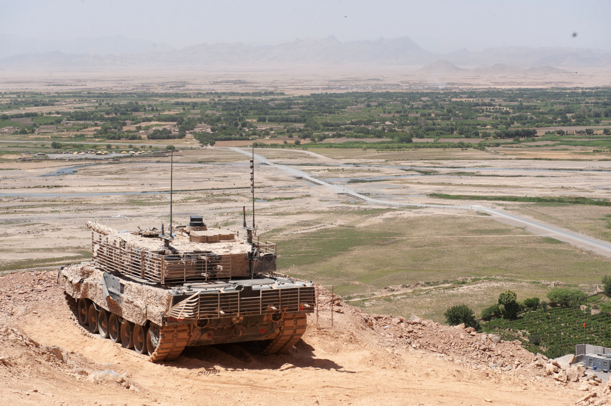 Un char de combat militaire de couleur beige camouflage trônant sur une hauteur qui surplombe un vaste paysage verdoyant et plat, avec une chaine de montagnes au loin