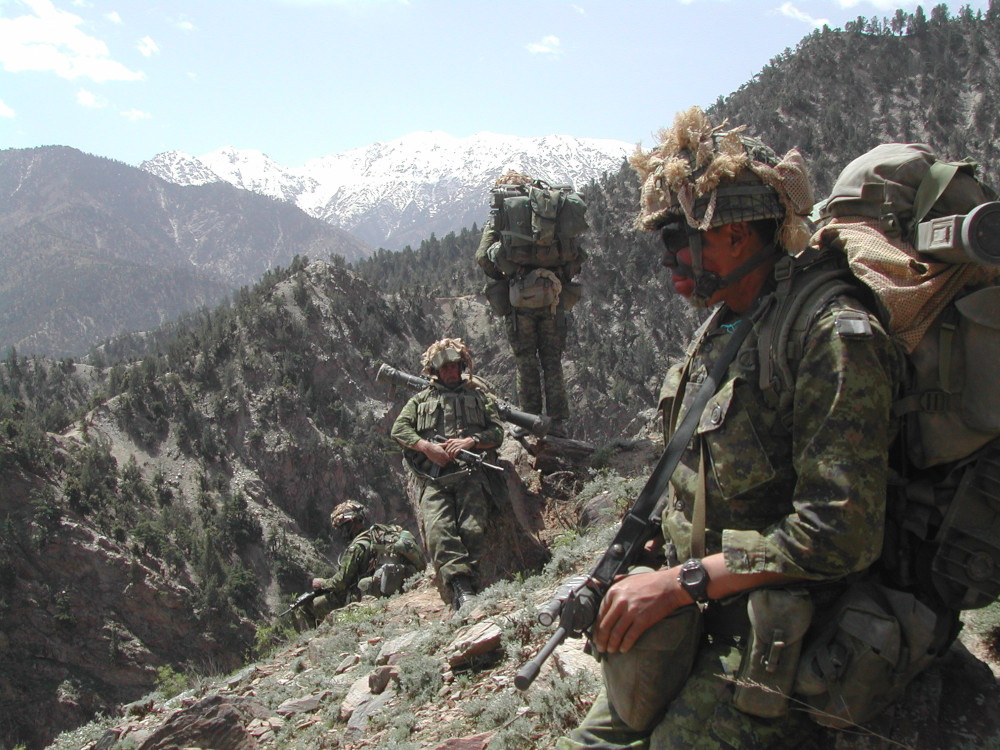 Quatre militaires en tenue de camouflage verte et au visage peint se tiennent sur une pente, avec, en arrière-plan, des crêtes montagneuses proches et lointaines.