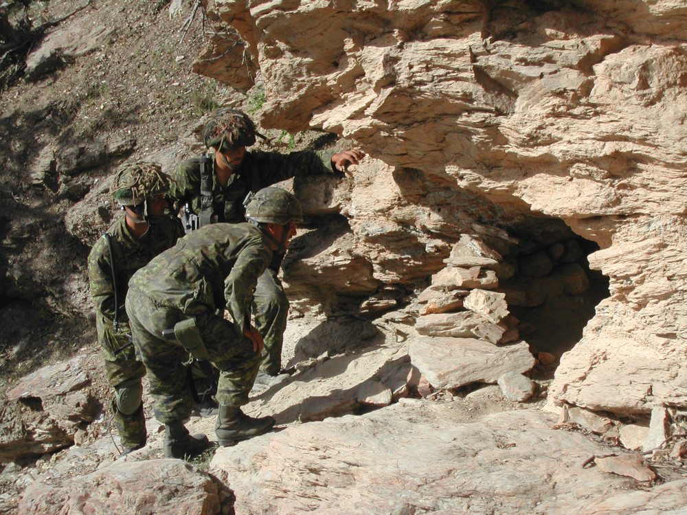 Trois militaires en tenue de camouflage verte scrutent l’entrée d’une petite grotte.