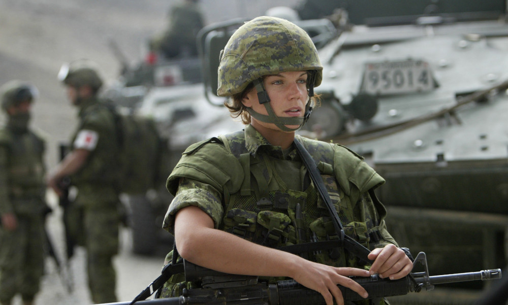 Une soldate vêtue d’une tenue de camouflage vert et d’un casque, une arme à la main, marche à côté de gros véhicules blindés; on aperçoit d’autres militaires derrière elle.