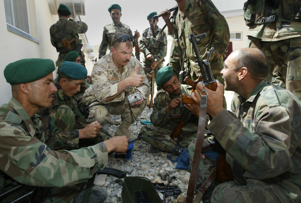 Un soldat à la peau pâle, portant une moustache et un uniforme de camouflage beige, est accroupi et lève le pouce au milieu d’un groupe de militaires à la peau moyennement foncée portant un uniforme de camouflage vert et tenant des mitraillettes ou s’affairant à les entretenir.