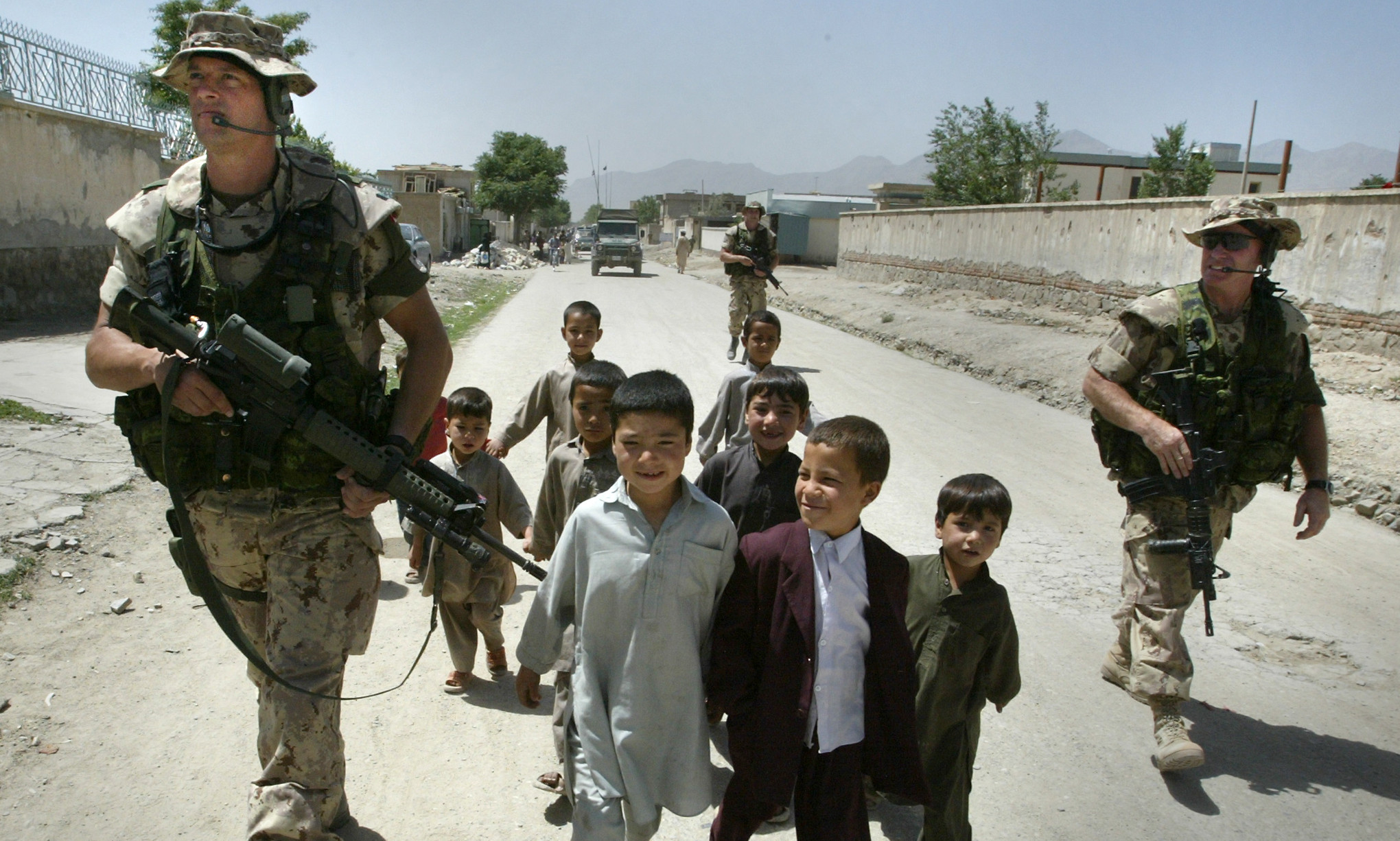 Trois militaires en tenue de camouflage beige, avec leur arme, marchent sur un chemin de terre bordé de murs de boue, en compagnie d’un groupe de jeunes enfants.