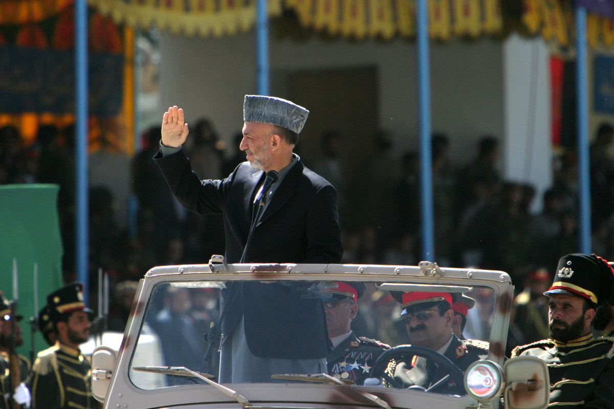 Un homme au teint moyen, portant une courte barbe grisonnante, un chapeau gris et un veston foncé, est debout dans une grande voiture décapotable et salue de la main, entouré d’hommes assis vêtus d’uniformes richement décorés.