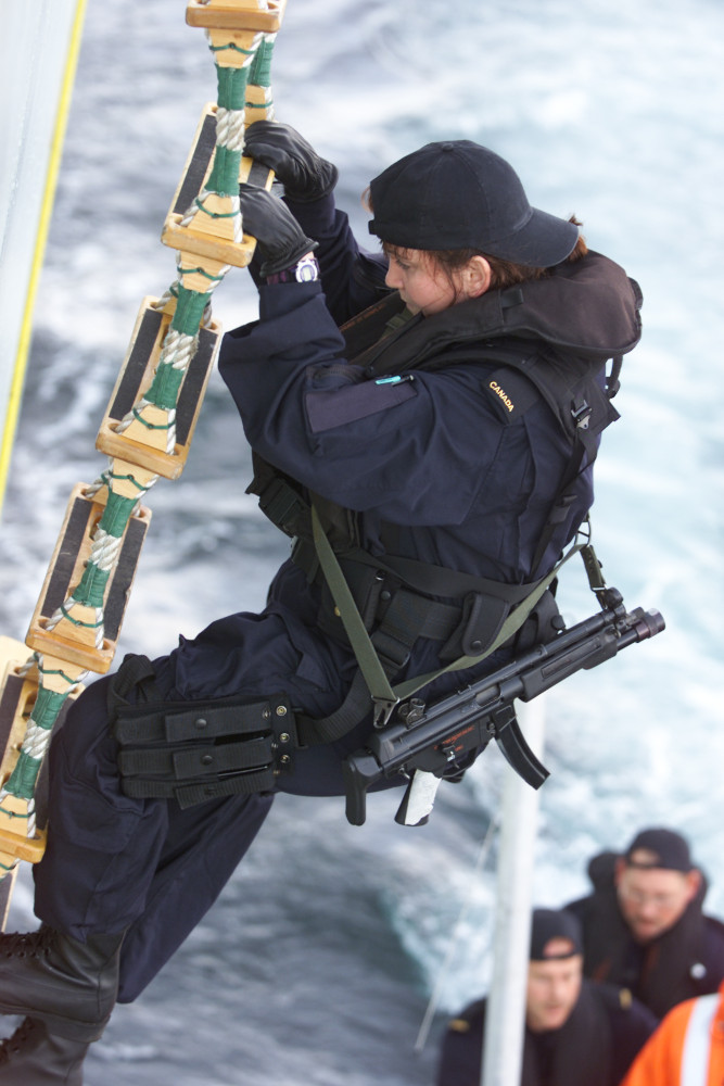 Une femme vêtue d’un uniforme bleu foncé et portant une mitraillette en bandoulière grimpe une échelle de corde raide au-dessus d’un plan d’eau; on aperçoit partiellement, en contrebas, des personnes dans un petit bateau.