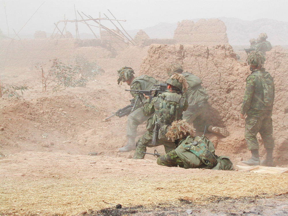 Un groupe de plusieurs militaires en tenue de camouflage verte, armes à la main, se protège derrière un mur de boue tout en scrutant les environs; on aperçoit au loin d’autres constructions en boue et en bois.