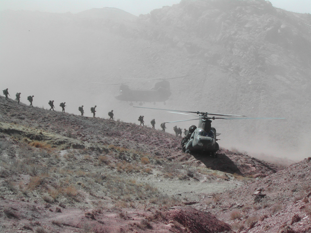 La silhouette d’une file de militaires marchant le long d’une crête en direction d’un grand hélicoptère, tandis qu’un autre hélicoptère demeure en vol stationnaire un peu plus loin; une montagne se profile à l’horizon.