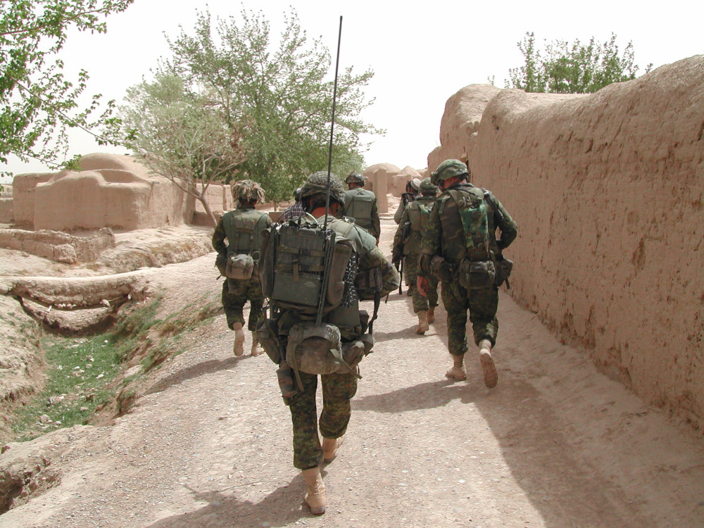 Un groupe de militaires en tenue de camouflage verte s’éloigne sur un chemin de terre, au milieu de murs et de bâtiments beiges.