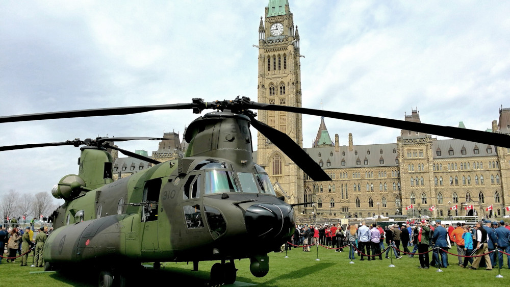 Un grand hélicoptère vert et une foule de gens devant les édifices du Parlement canadien.