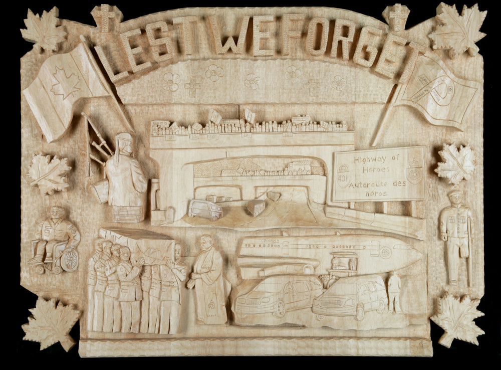 Une sculpture de bois en relief avec « Lest we forget (N’oublions jamais) » en grandes lettres dans la partie supérieure, et des scènes du rapatriement d’une dépouille de militaire, y compris un groupe de militaires qui portent un cercueil et une foule rassemblée sur un viaduc d’autoroute.