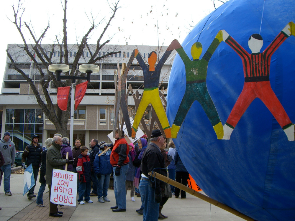 Un groupe de personnes est rassemblé sur une place publique. Au premier plan, on aperçoit une grande sculpture représentant un globe terrestre entouré de silhouettes humaines entrelacées.